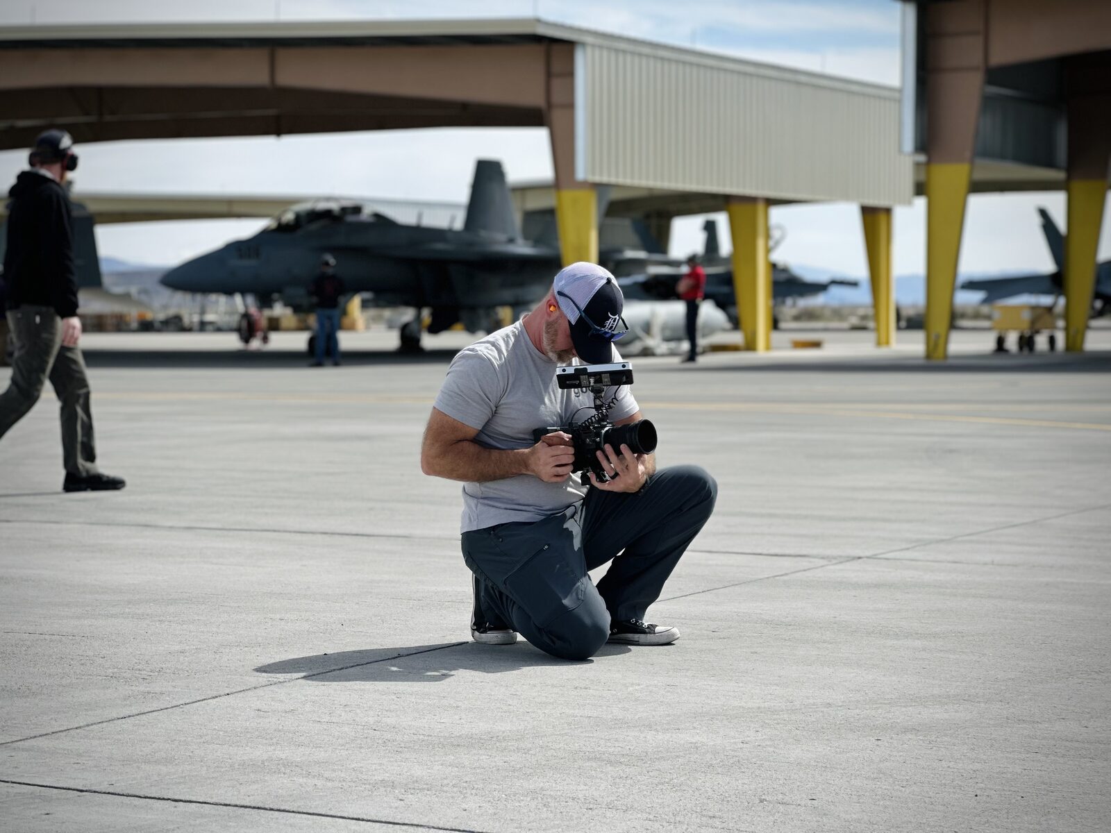 Camera operator kneeling with a cinema rig on an airfield tarmac with fighter jets behind, behind the scenes of Mind Shift Fights On production