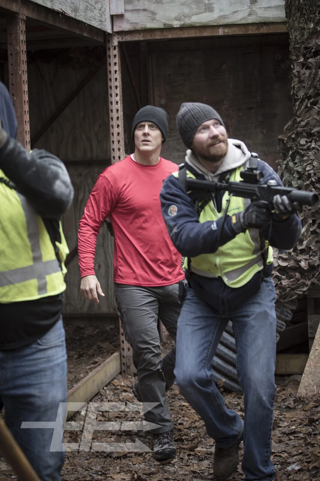 Two men in high-visibility vests advance through a rusted structure during a field training exercise, one aiming a submachine gun, tactical photography by Mind Shift