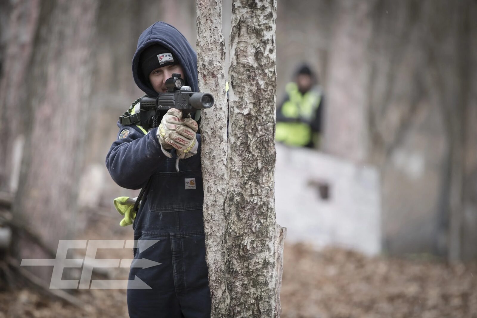 Hooded participant aims a scoped rifle from behind a tree while an observer watches during a field training exercise, tactical photography by Mind Shift