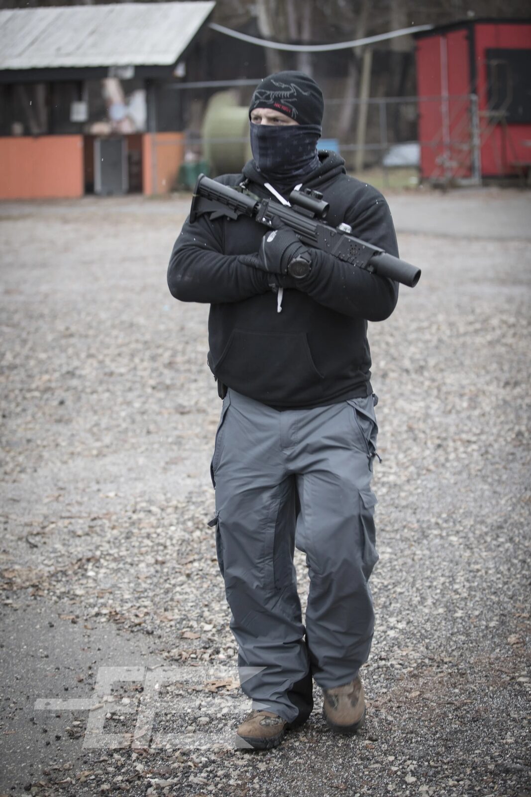 Masked participant in tactical gear carries a scoped rifle across a gravel lot during a field training exercise, tactical photography by Mind Shift